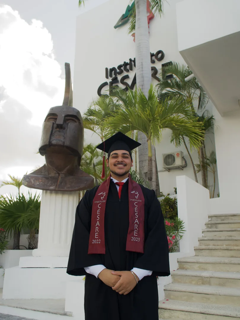 Graduado con toga y birrete sonríe junto a la escultura del casco de César.