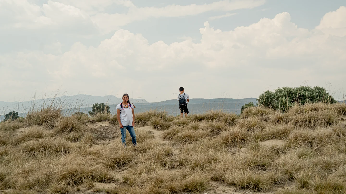 Mujer y niño separados sobre un cerro de pasto seco bajo un cielo nublado.