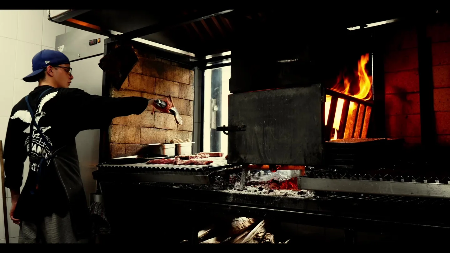 Cocinero de espaldas con gorra azul atiende la parrilla junto a un horno con fuego.