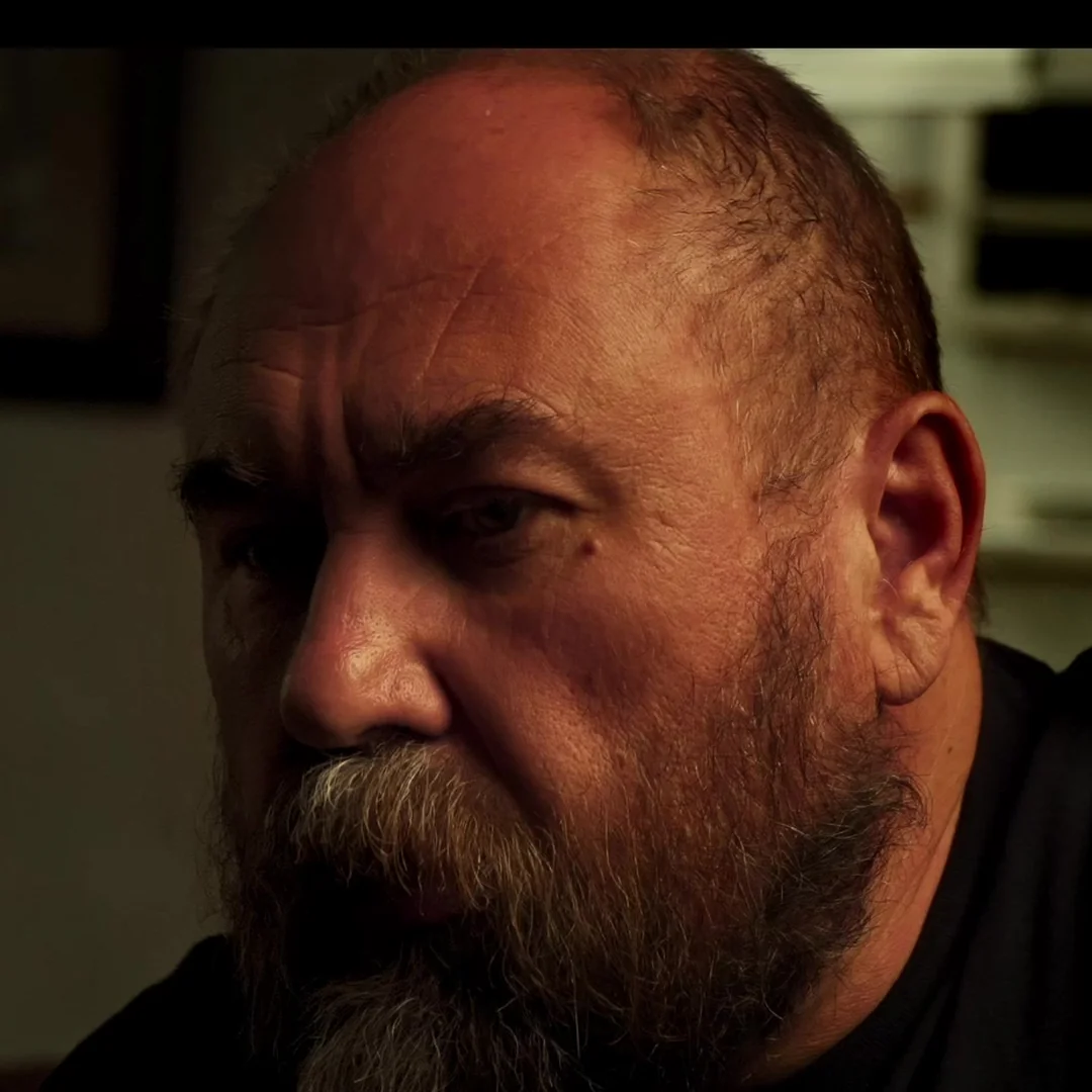 Primer plano de un hombre calvo con barba canosa, iluminado de lado sobre fondo oscuro.